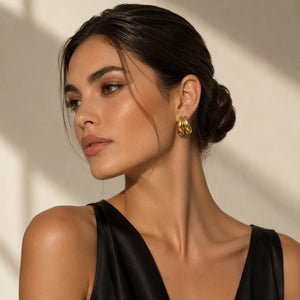 A close-up of a woman with dark hair in a bun wearing a pair of chunky, interlocking gold hoop earrings, set against a warm beige wall with soft diagonal shadows.