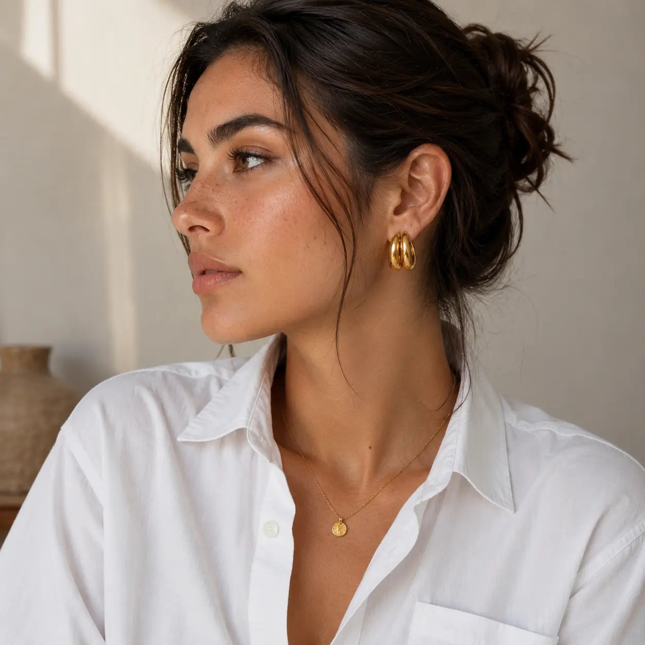 A woman with dark hair in a bun wearing a white button-up shirt, showcasing a pair of chunky gold hoop earrings and a delicate gold necklace with a small disc pendant.