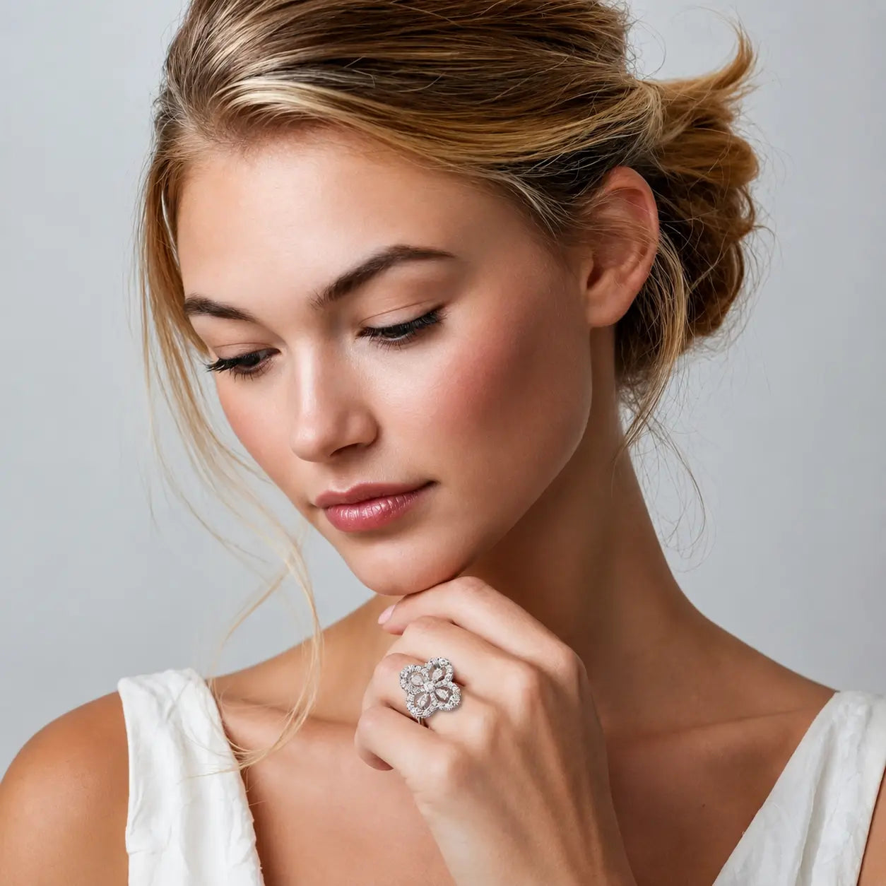 A close-up lifestyle shot of a woman with blonde hair in an updo, wearing a large, intricate silver ring with a flower-like design set with clear stones. She is wearing a white top against a neutral gray background.