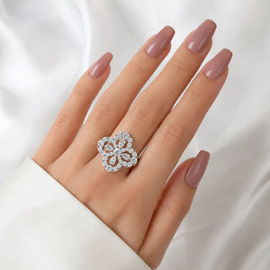 A silver-toned flower ring with clear stones is worn on a hand with long, manicured nude-pink nails, set against a soft white satin background.