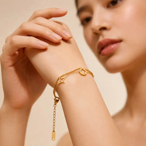 A close-up lifestyle shot of a model wearing a delicate gold chain bracelet on her wrist. The bracelet features a star-shaped charm, a circular medallion charm, and a dangling tag charm, set against a soft beige background.