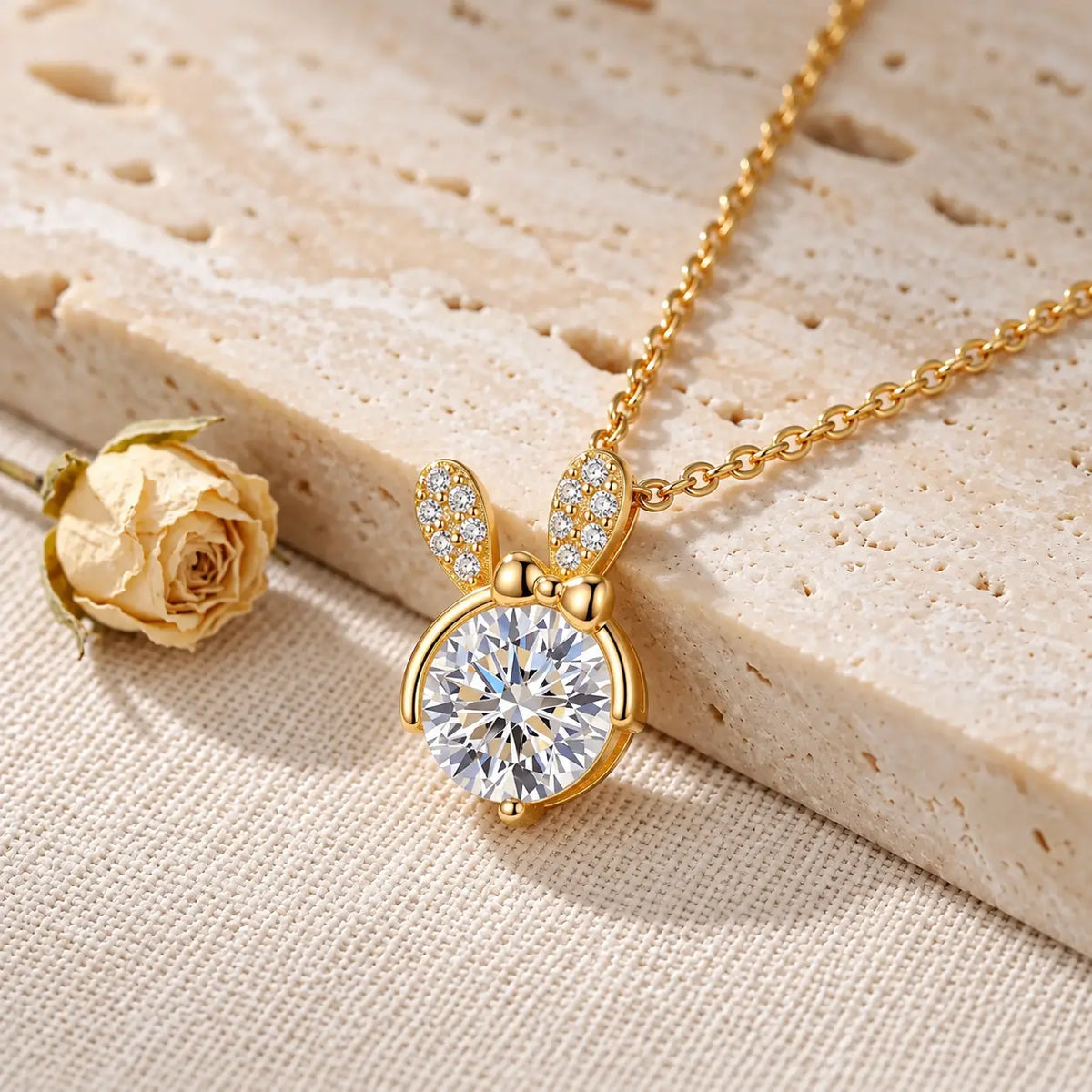 Gold bunny pendant necklace with a large clear round gemstone and small clear stones on the ears, displayed on a textured beige surface next to a dried rose.