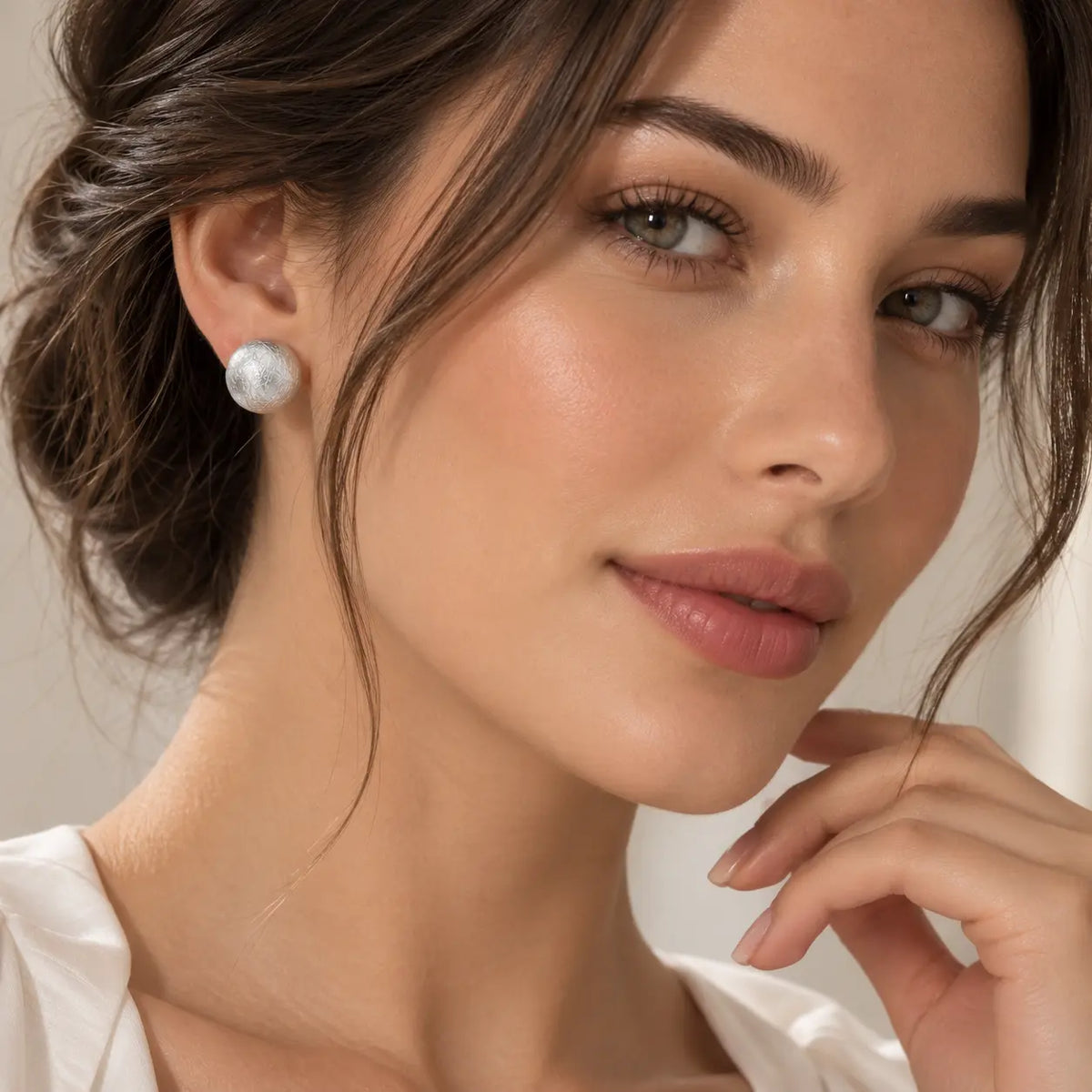 A close-up portrait of a woman wearing a single round white pearl stud earring. The earring is positioned on her ear, with her hand resting near her chin.