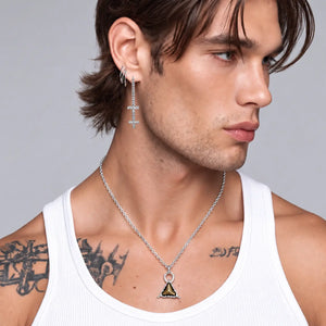 A close-up of a man wearing a silver-toned geometric pendant necklace and matching drop earrings, paired with a white ribbed tank top against a neutral gray background.