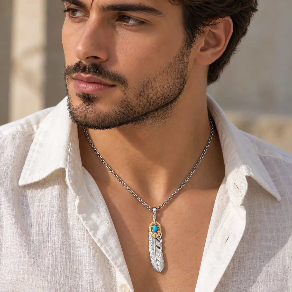A close-up of a man wearing a silver-toned chain necklace with a feather pendant featuring a turquoise blue stone and gold accents, paired with an open white linen shirt.
