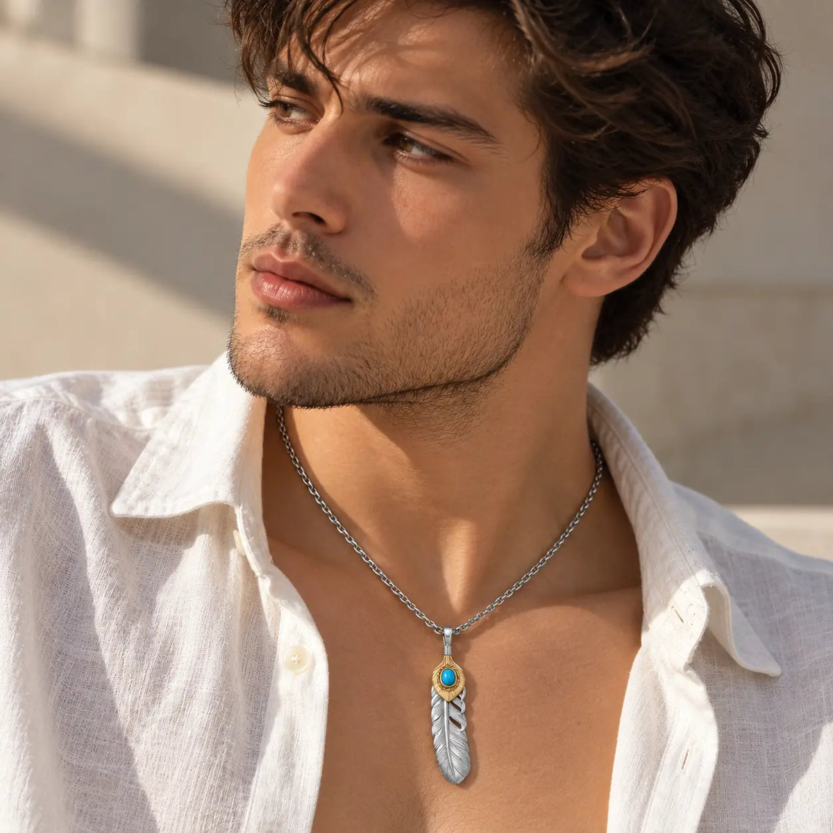A man wearing a silver-toned feather pendant necklace with a turquoise accent, styled with an open white linen shirt.