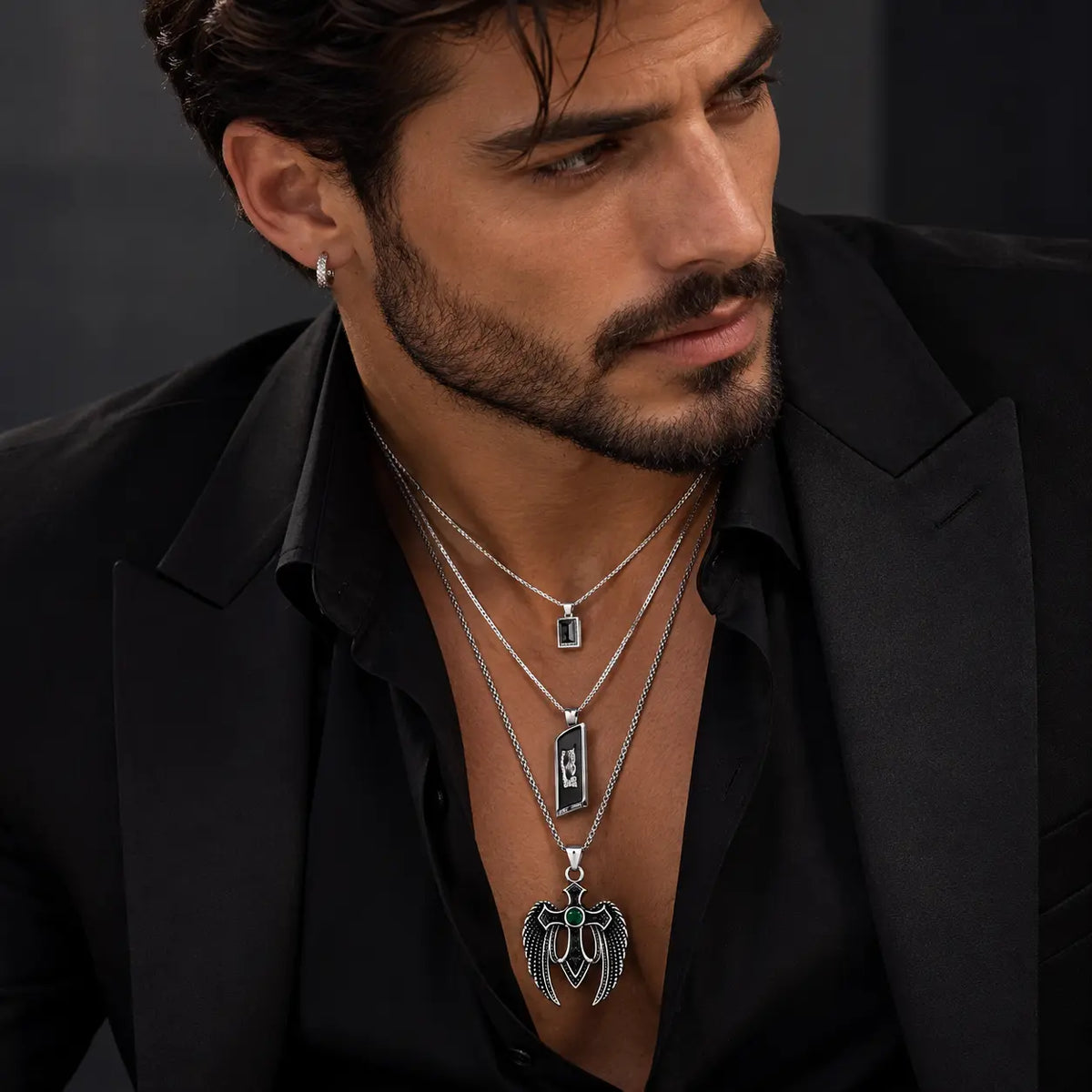 A close-up of a man wearing layered silver necklaces with rectangular and winged pendants over a black shirt and blazer.