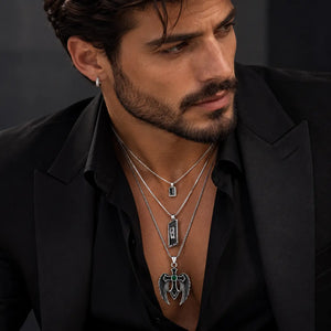 A close-up of a man wearing layered silver necklaces with rectangular and winged pendants over a black shirt and blazer.