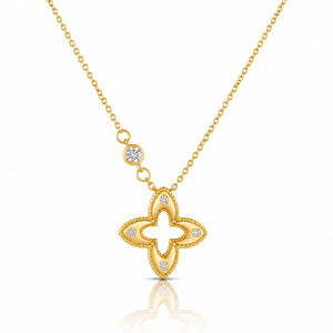 A gold necklace featuring a four-lobed clover or quatrefoil pendant with small clear gemstones set along the edges, suspended from a delicate gold chain with a small round stone accent, shown on a white background with a soft reflection below.