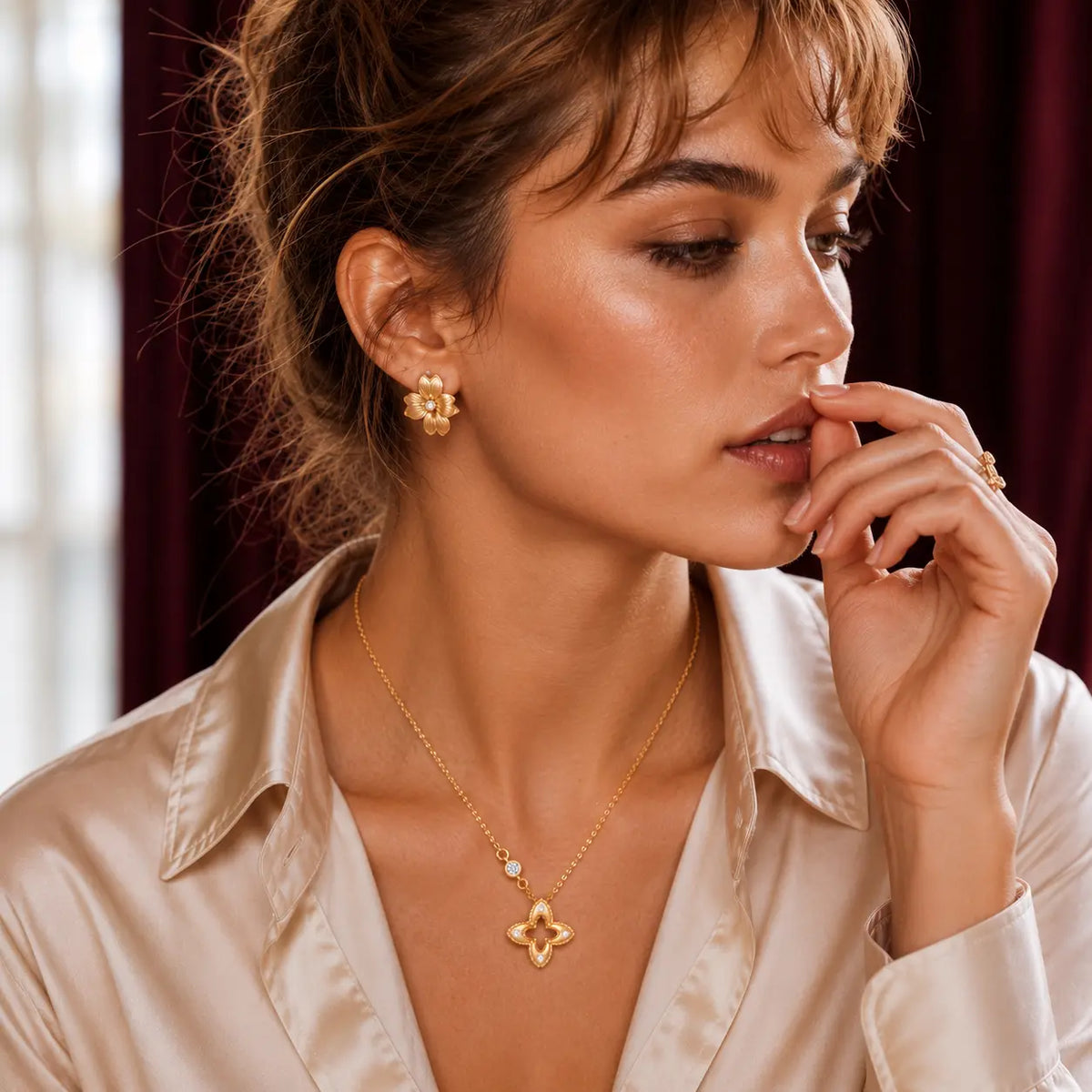A close-up portrait of a model wearing gold jewelry, including a gold necklace with a clover-shaped pendant, a matching ring, and earrings, set against a dark red curtain background.