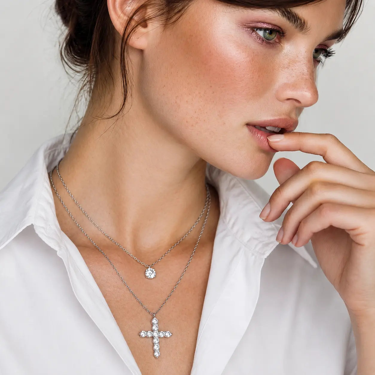 A close-up lifestyle shot of a woman wearing two layered silver necklaces: a delicate chain with a small round clear gemstone pendant and a longer chain with a silver cross pendant set with clear stones, styled with a white collared shirt.