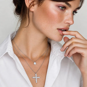 A close-up lifestyle shot of a woman wearing two layered silver necklaces: a delicate chain with a small round clear gemstone pendant and a longer chain with a silver cross pendant set with clear stones, styled with a white collared shirt.