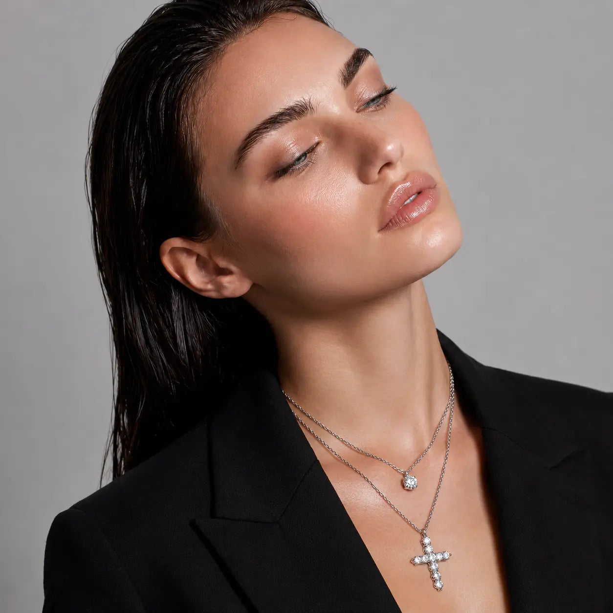A close-up of a woman wearing two silver-toned necklaces, including a cross pendant and a small round pendant, styled with a black blazer against a neutral gray background.