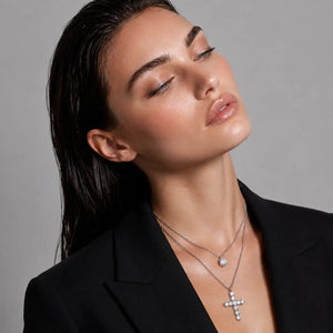 A close-up of a woman wearing two silver-toned necklaces, including a cross pendant and a small round pendant, styled with a black blazer against a neutral gray background.