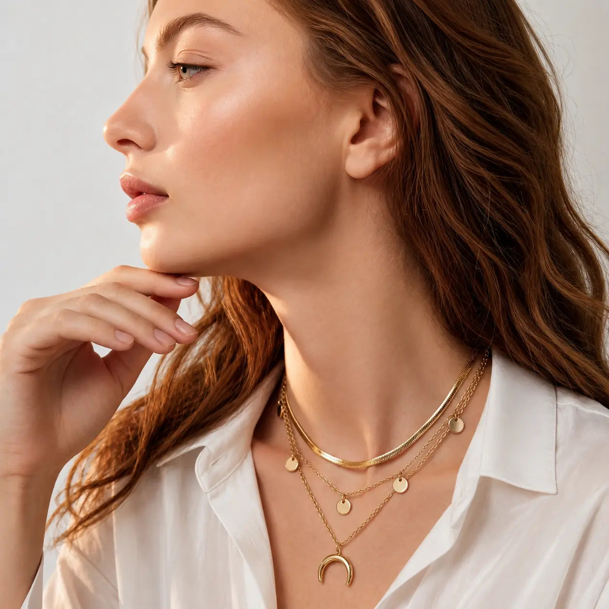 A close-up of a woman wearing layered gold necklaces, including a bangle, delicate chain with disc charms, and a crescent moon pendant, paired with a white collared shirt.