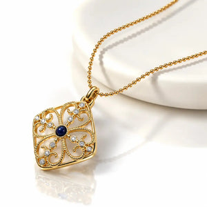A gold filigree pendant necklace with a central dark blue gemstone and small clear rhinestones, displayed on a white curved stand against a white background.