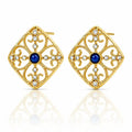 A pair of gold filigree stud earrings with a central deep blue gemstone and small clear rhinestones, displayed on a clean white background with a soft reflection below.