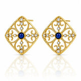 A pair of gold filigree stud earrings with a central deep blue gemstone and small clear rhinestones, displayed on a clean white background with a soft reflection below.