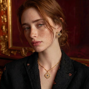 Woman wearing gold jewelry against a richly decorated wall.