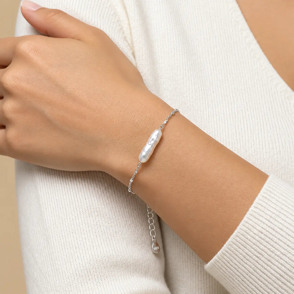 A close-up of a person's wrist wearing a delicate silver chain bracelet with a single baroque pearl pendant, paired with a cream-colored knit sweater.