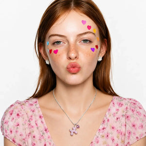 A young woman with freckles wearing a pink floral top, a silver necklace with a butterfly pendant, and small heart-shaped face gems on her forehead and cheeks, posing with a puckered expression against a plain white background.