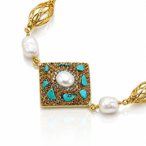 A gold-tone necklace featuring a square pendant with turquoise and brown pavé accents, a central white pearl, and two baroque pearls on a white background.