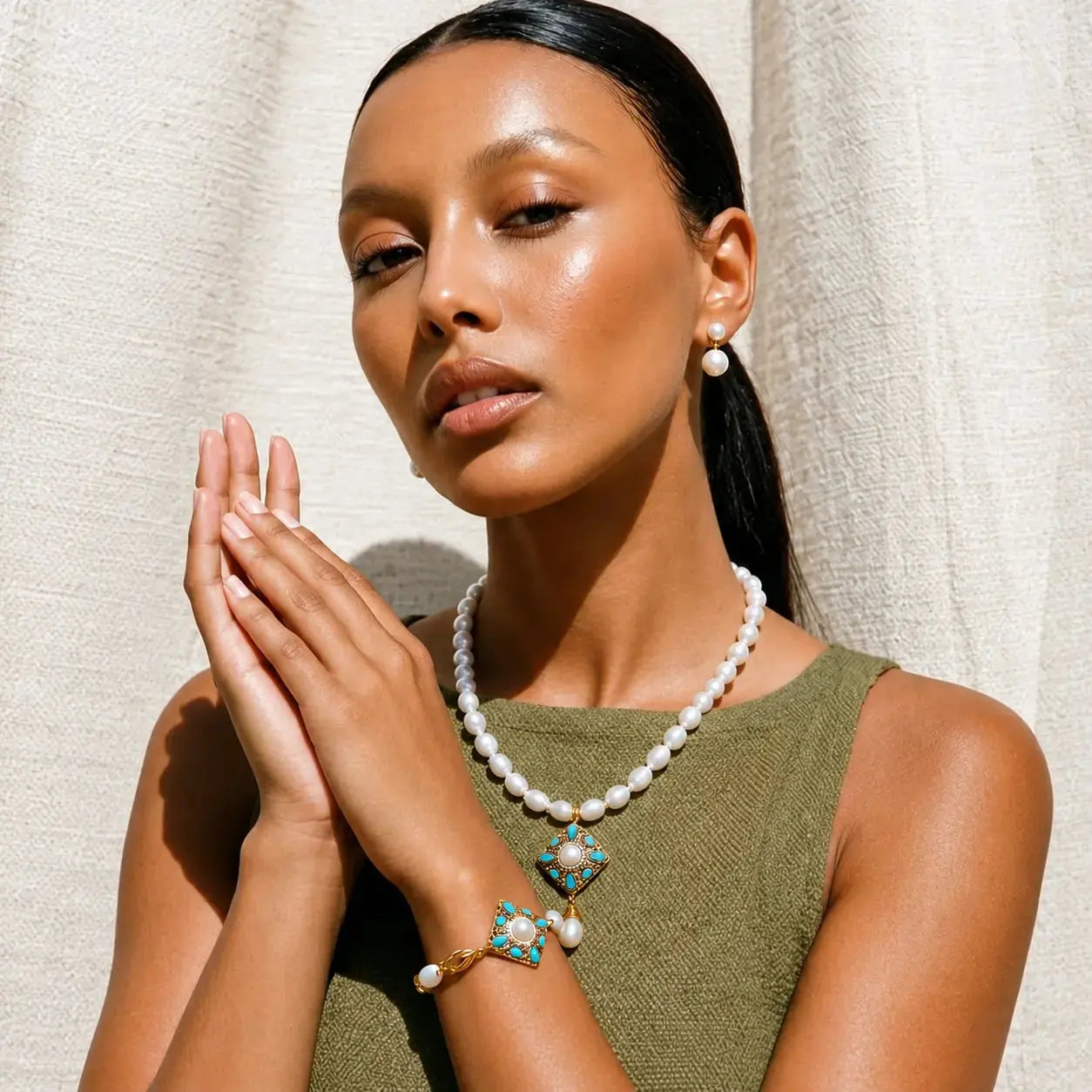 A woman with her hair in a ponytail models a white pearl necklace and matching pearl earrings with turquoise and gold accents, paired with an olive green top against a textured cream curtain background.