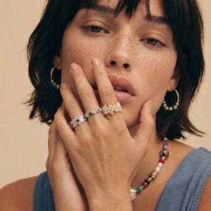 Woman wearing gold hoop earrings, a ring, and a colorful necklace against a beige background