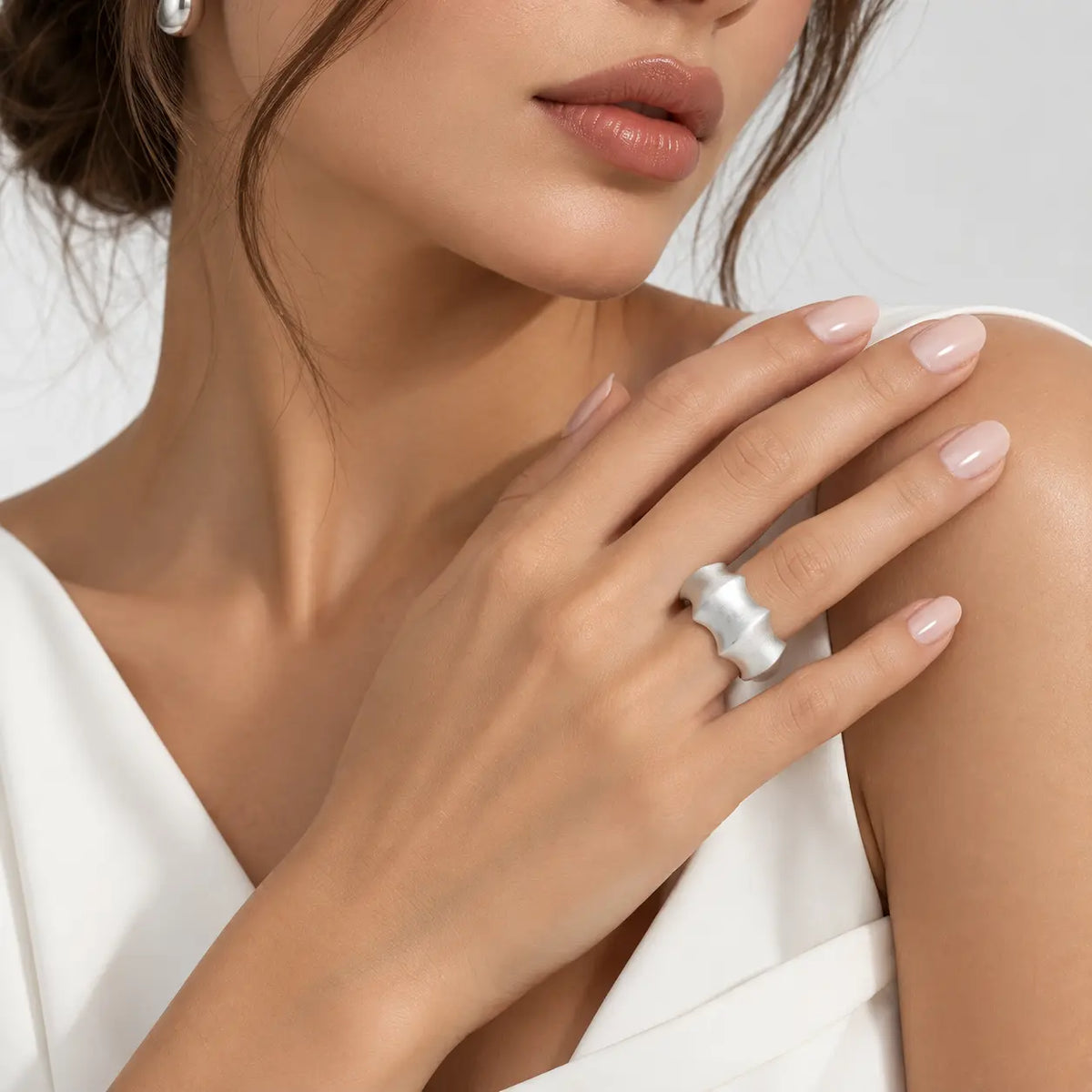 A close-up lifestyle shot of a woman wearing a wide, chunky silver ring on her middle finger, paired with a white top against a soft light gray background.