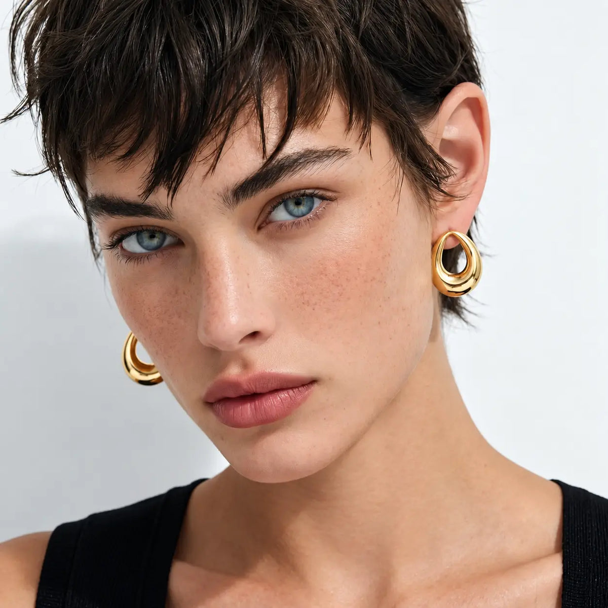 A close-up portrait of a model with short dark hair wearing gold hoop earrings with dark stone accents, against a clean light gray background.
