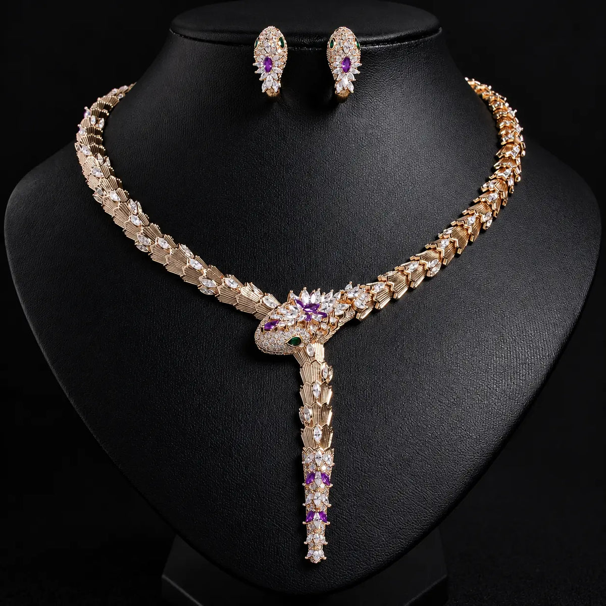 Gold-tone statement necklace and matching stud earrings displayed on a black jewelry bust. The necklace features clear rhinestones, a central purple gemstone, and a vertical drop with purple and green gemstone accents.