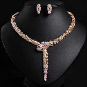 Gold-tone statement necklace and matching stud earrings displayed on a black jewelry bust. The necklace features clear rhinestones, a central purple gemstone, and a vertical drop with purple and green gemstone accents.