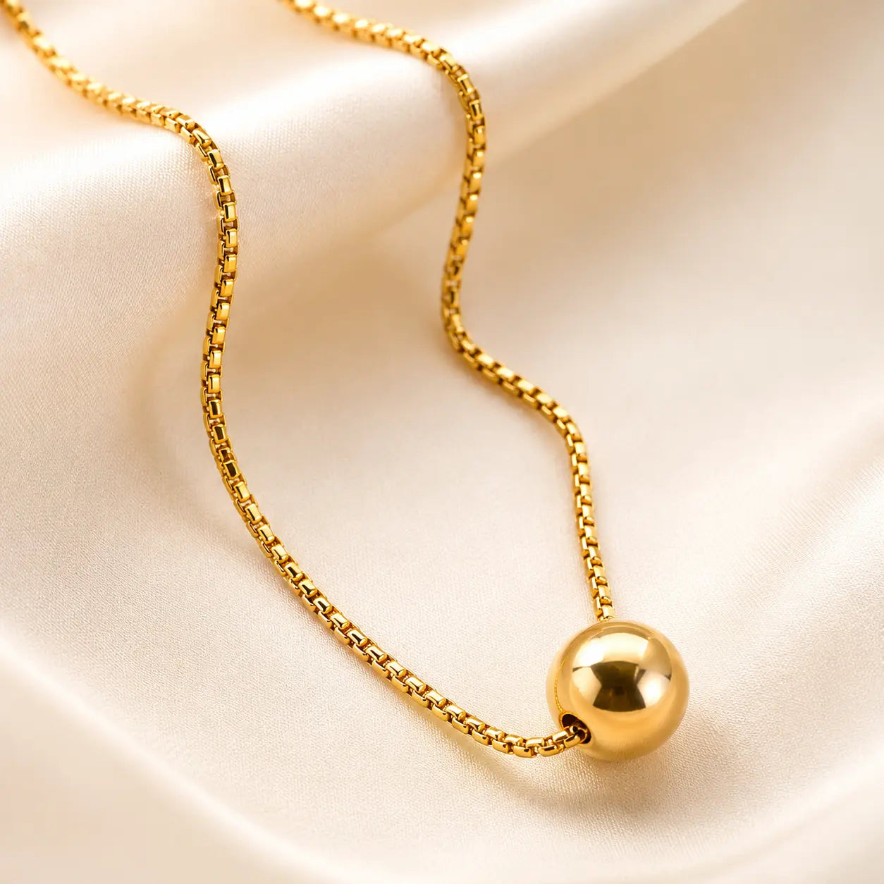 A gold chain necklace with a polished spherical pendant, displayed on draped cream satin fabric.