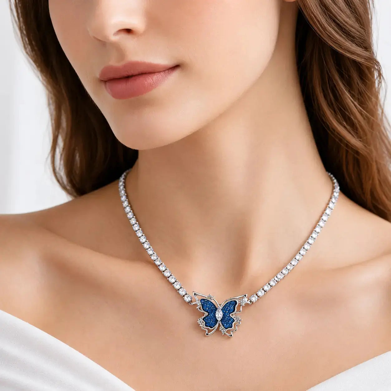 A close-up of a woman wearing a delicate silver-toned necklace with a butterfly pendant. The butterfly has vibrant blue enamel inlay and is set on a silver-toned chain studded with small clear crystals.