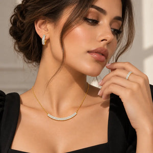A close-up lifestyle shot of a woman wearing a gold necklace with a curved row of white pearls, a matching pearl hoop earring, and a pearl ring on her finger, set against a soft beige background.