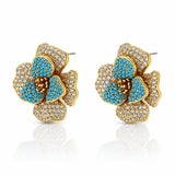 A pair of gold flower-shaped stud earrings adorned with turquoise blue pavé stones and clear crystal-like accents, displayed on a clean white background with a soft reflection below.