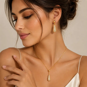 A woman wearing a gold teardrop pendant necklace and matching drop earring, both adorned with sparkling clear stones, against a soft beige background.