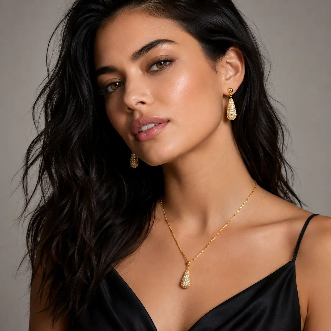 A woman with long dark wavy hair models a gold teardrop pendant necklace and matching drop earrings with sparkling clear stones against a neutral gray background.