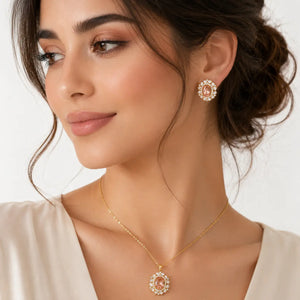 A close-up of a woman wearing a gold necklace and matching stud earring with a peach-pink and white floral design, set against a clean white background.
