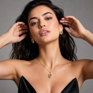 A close-up portrait of a woman wearing a delicate gold necklace with a circular pendant and matching stud earrings, styled with a black satin top against a neutral gray background.