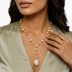 A close-up of a woman wearing a gold and pearl necklace with a large teardrop pearl pendant, paired with an olive green satin top.