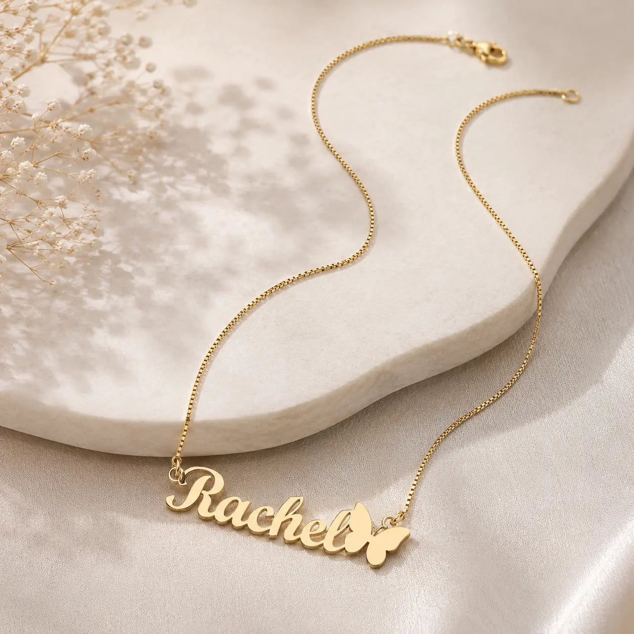 A gold nameplate necklace spelling 'Rachel' with a butterfly accent, displayed on a cream-colored stone slab with soft shadows.