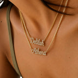 Close-up of a person wearing two layered gold necklaces with script name pendants, one reading 'Deblah' and the other 'Athena', set with small clear stones.