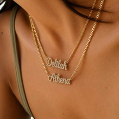 Close-up of a person wearing two layered gold necklaces with script name pendants, one reading 'Deblah' and the other 'Athena', set with small clear stones.