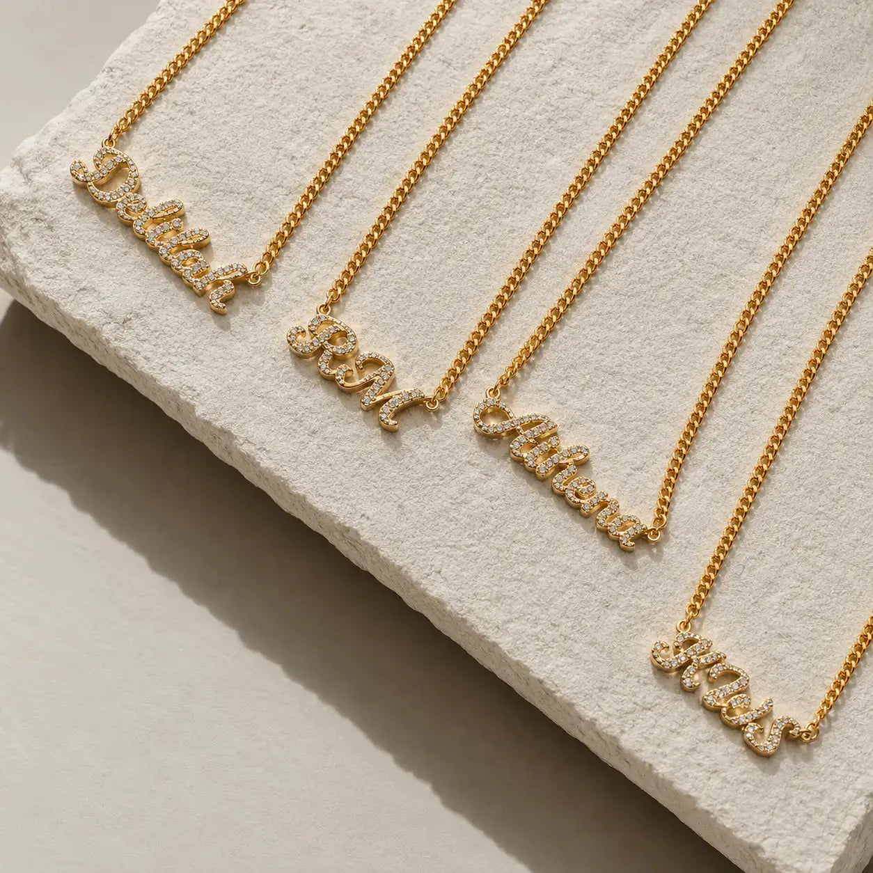 Four gold nameplates on matching chains arranged diagonally on a textured white display block. The nameplates are personalized with words like 'Daddy', 'Mom', 'Father', and 'Miss' in a script style with small clear stones.