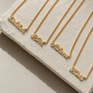 Four gold nameplates on matching chains arranged diagonally on a textured white display block. The nameplates are personalized with words like 'Daddy', 'Mom', 'Father', and 'Miss' in a script style with small clear stones.