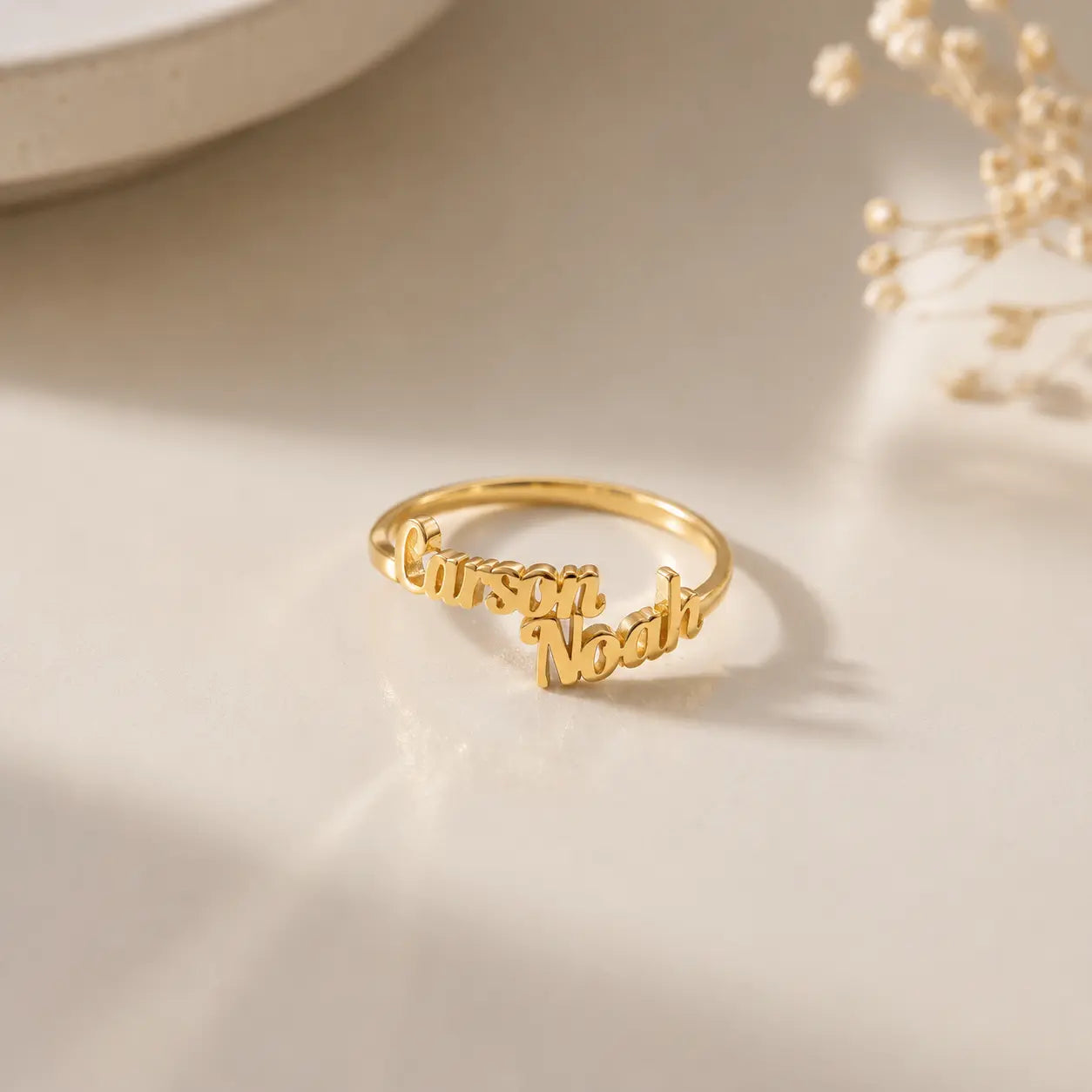 A gold name ring engraved with the names 'Cursari' and 'Noah' in cursive script, displayed on a light beige surface with soft shadows and dried flowers in the background.
