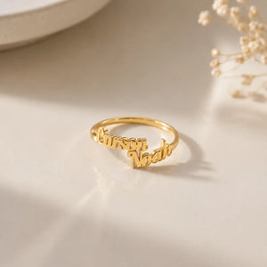 A gold name ring engraved with the names 'Cursari' and 'Noah' in cursive script, displayed on a light beige surface with soft shadows and dried flowers in the background.