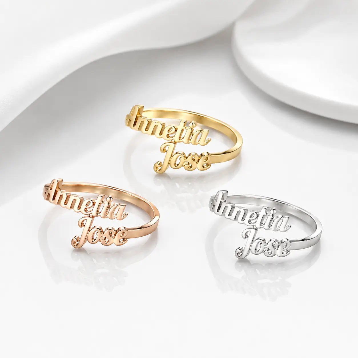 Three personalized name rings reading 'Annetta Jose' in gold, rose gold, and silver, arranged on a clean white reflective surface with soft draped fabric in the background.