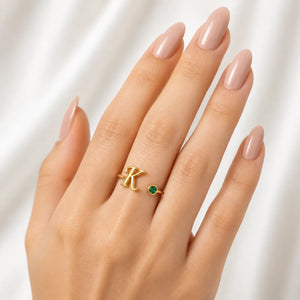 A close-up of a hand with manicured nails wearing a gold ring featuring a large letter 'K' and a small round green stone.